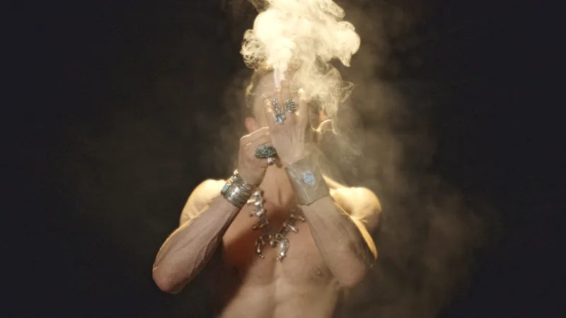 Still from the Dancing Art Dealer pitch teaser: a shirtless man in turquoise jewelry covers his face as smoke rises